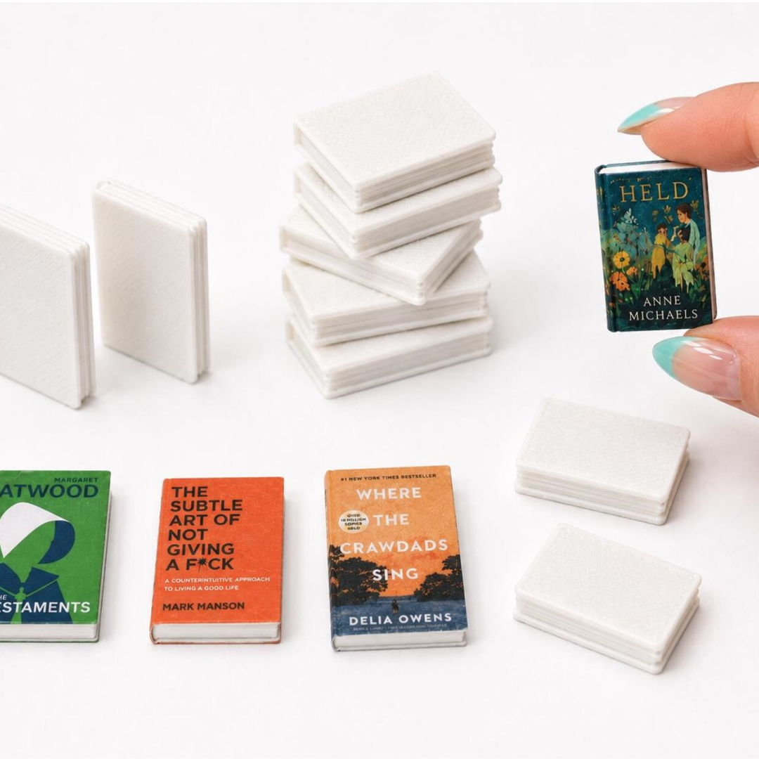 A set of miniature book-shaped items, including several stacks of plain white book covers and three printed book covers, displayed on a white background with a hand holding one book.