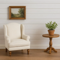 An off-white upholstered wingback armchair sits beside a small round wooden side table holding a potted green plant, against a white shiplap wall with a framed landscape painting above.